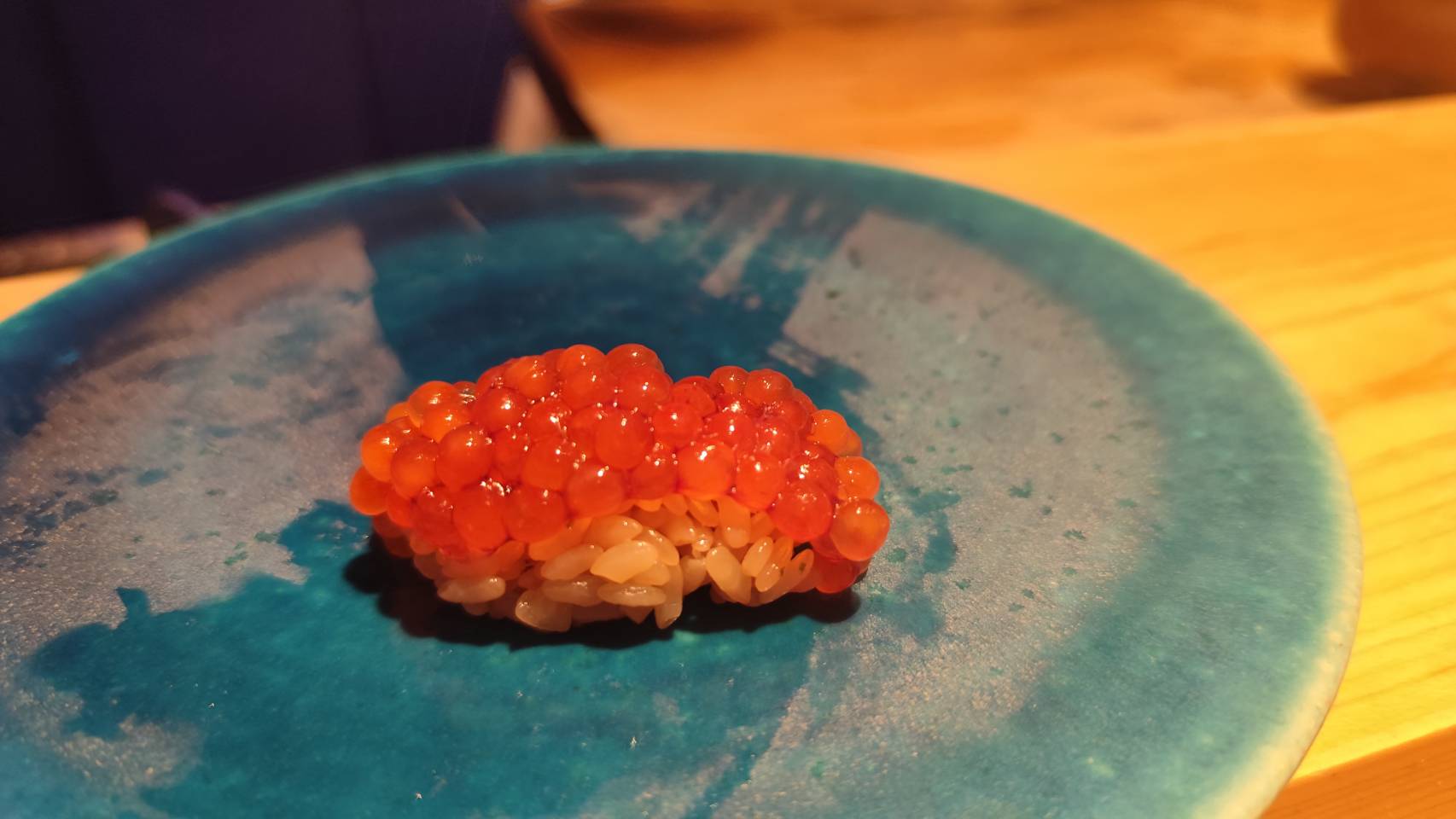 青い皿に盛られたイクラ寿司。輝くオレンジ色の粒が美しく並び、酢飯にのせられた贅沢な一皿。