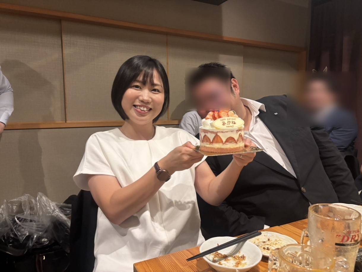 誕生日ケーキを持ちながら笑顔を見せる女性と、青年会議所の仲間たちに囲まれている様子。