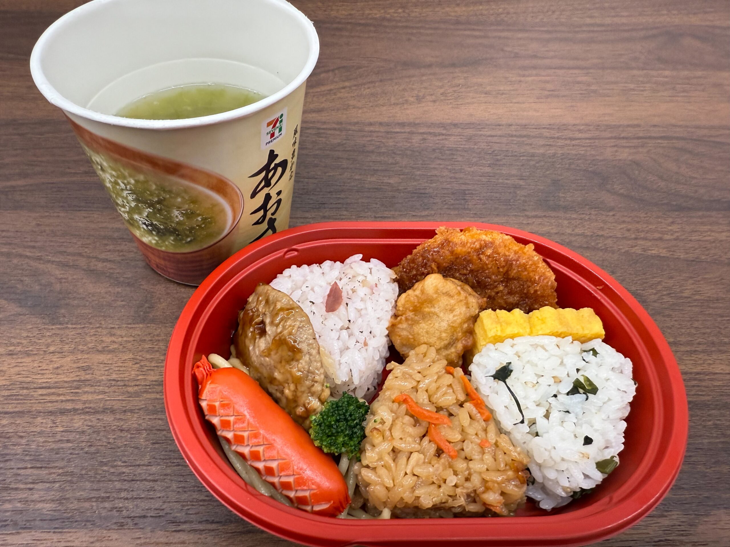 セブンイレブンのお弁当とあおさ味噌汁。おにぎり3種類と卵焼き、コロッケ、唐揚げ、ウィンナーが彩りよく盛られている。
