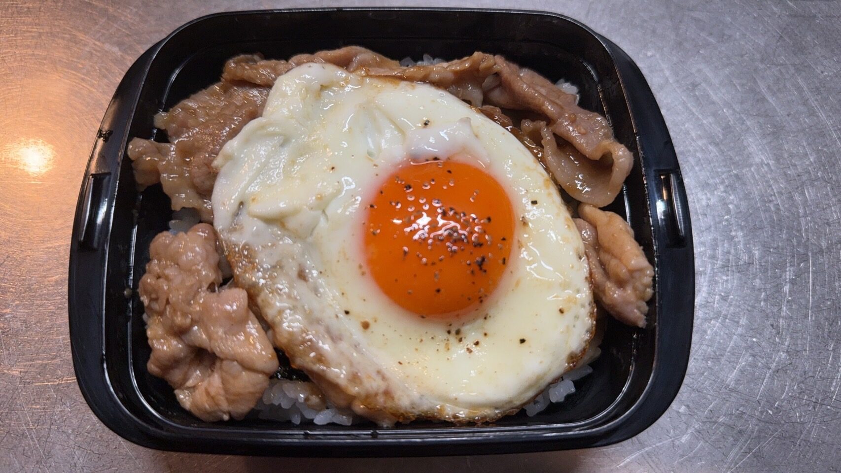 黒い弁当容器に盛られた自分用の豚の焼肉弁当。ご飯の上に焼肉と目玉焼きがのり、スタミナ満点で美味しそう。