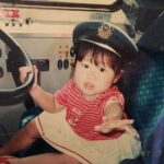 3歳の頃、バスの運転席に座り運転手帽をかぶっている幼い女の子の写真。