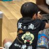 「飯と肉。」のオリジナルTシャツを着た子ども。背中には力強いキャラクターと店名ロゴが入っている。