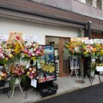 オープン当日にお祝いの花が並ぶ「飯と肉。」店舗前の写真。爆盛弁当＆ベーカリー晃蘭の看板も設置されている。