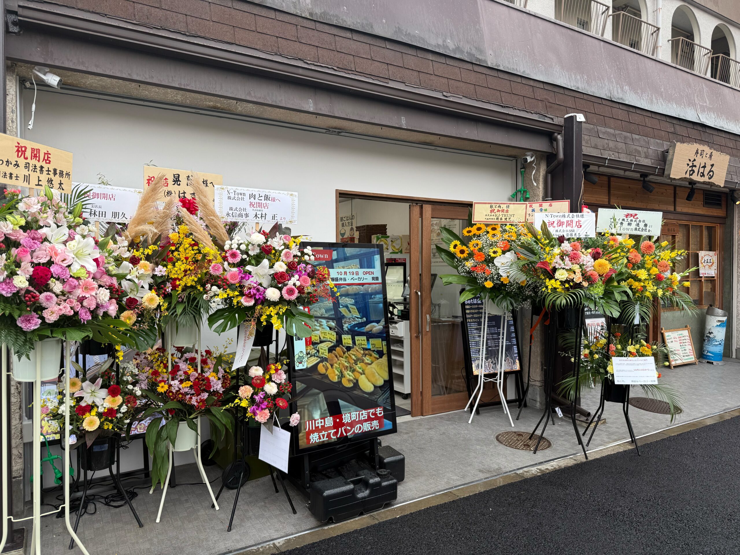 オープン当日にお祝いの花が並ぶ「飯と肉。」店舗前の写真。爆盛弁当&ベーカリー晃蘭の看板も設置されている。