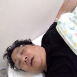 点滴を受けながら病院のベッドで休む男性。笑顔で「リゲイン」を歌っている様子が浮かぶ。