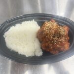 黒い楕円形の容器に白ご飯と白ごまがかかったヤンニョムチキンが盛られた弁当の写真