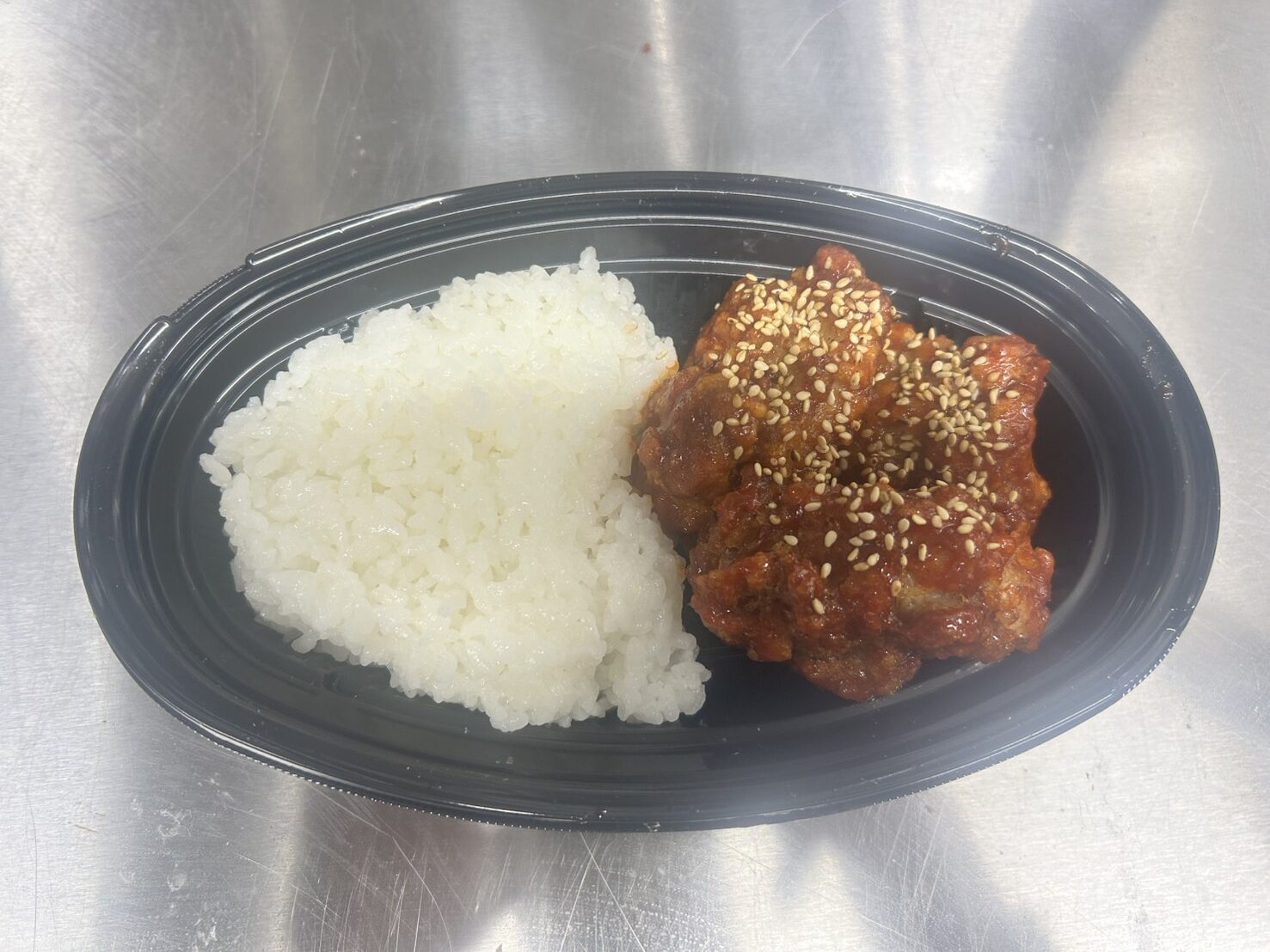 黒い楕円形の容器に白ご飯と白ごまがかかったヤンニョムチキンが盛られた弁当の写真