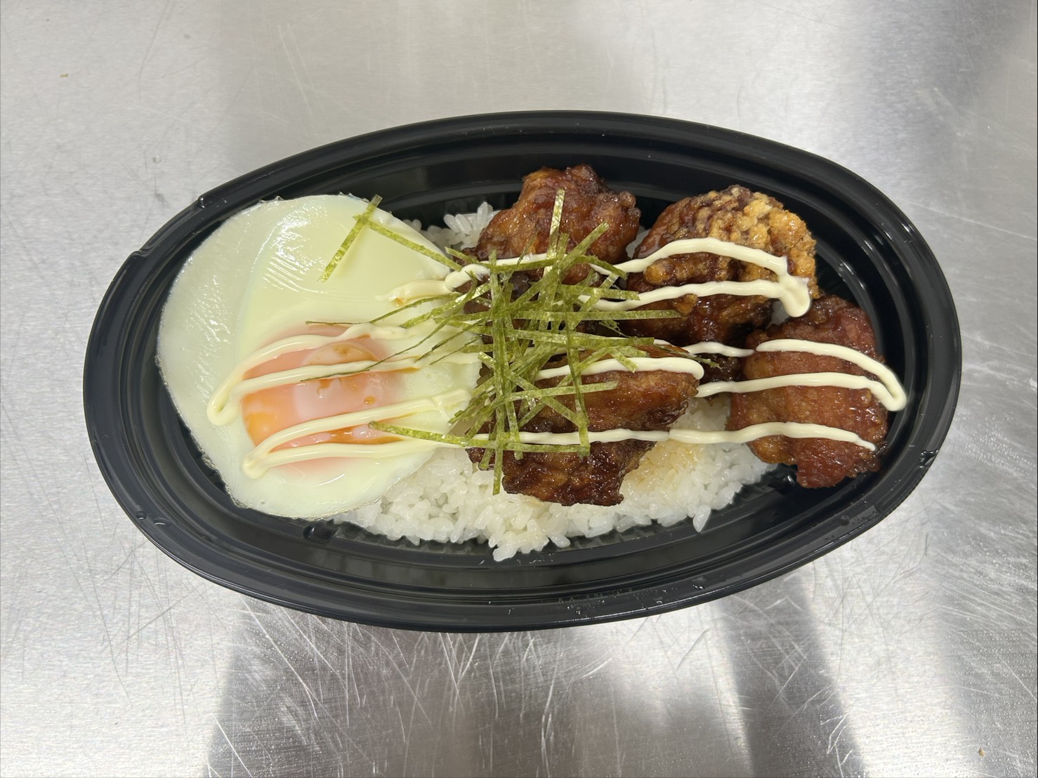 黒い楕円形の容器にご飯、その上に甘辛ダレとマヨネーズをかけた唐揚げと半熟目玉焼き、刻み海苔がのったマヨ唐丼の写真